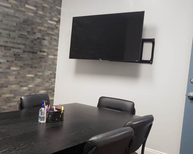 A conference room features a black table surrounded by six chairs, a wall-mounted television, and a stone-patterned accent wall.