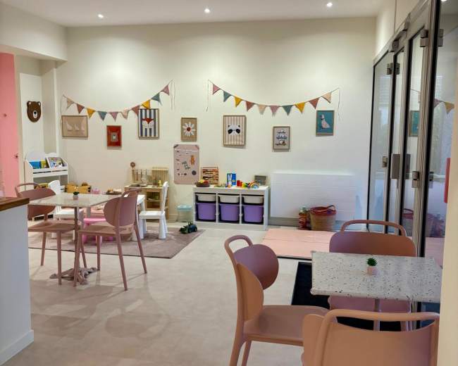 The image shows a modern playroom with tables, chairs, and colorful wall decorations, featuring toys and activity areas arranged neatly.
