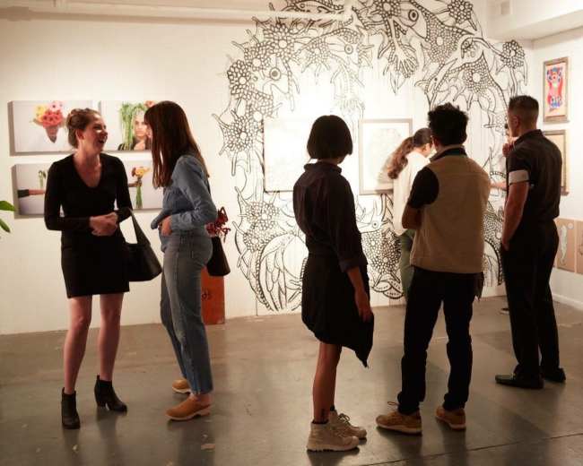 A group of people examines artwork in an art gallery, surrounded by various framed pieces and a mural on the wall.