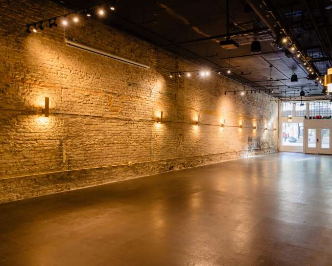 The image shows a spacious, well-lit interior with exposed brick walls and a polished floor, featuring minimal furnishings and a pair of double doors at the far end.