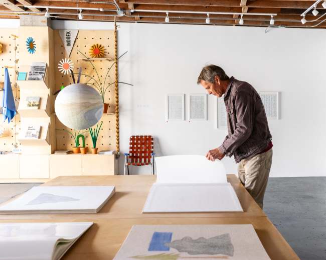A man stands at a table flipping through large sheets of paper in a brightly lit art studio with various artworks displayed on the walls.