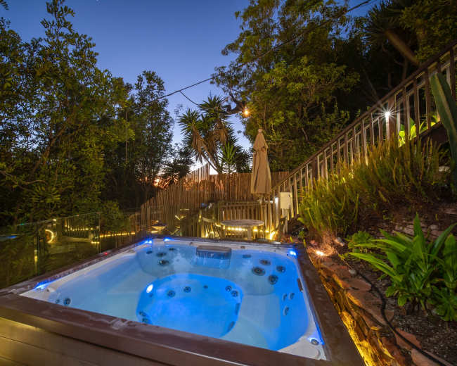 A hot tub is illuminated by blue lights in a landscaped outdoor area surrounded by trees and a staircase leading up to a deck.