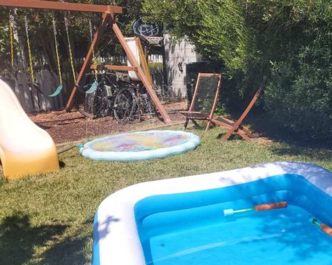 A small inflatable pool sits in a backyard next to a water slide and a playground swing set.