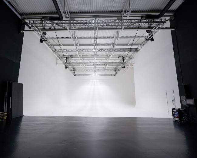 The image shows a spacious, empty studio with white walls and a polished black floor, illuminated by overhead lights.