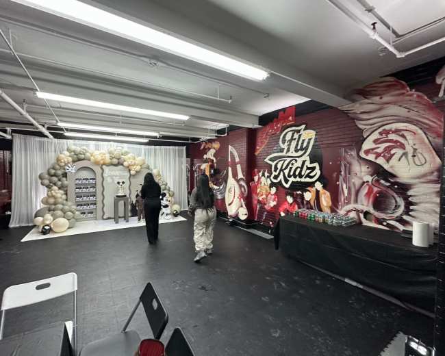The image shows a decorated indoor event space featuring a balloon arch, a display area, and a mural with the words "Fly Kidz."