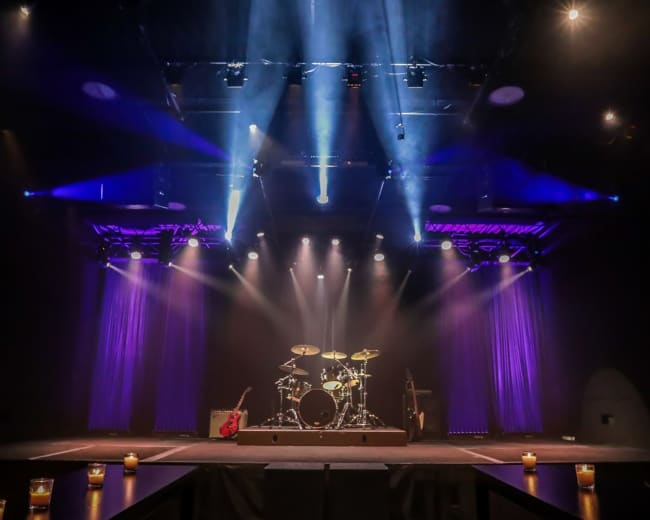 A stage is set with a drum kit and electric guitar, illuminated by blue and purple stage lights, surrounded by candlelit tables.