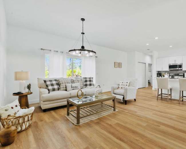 The image shows a modern living area with a light-colored sofa, a glass coffee table, and an open kitchen featuring white cabinetry and stainless-steel appliances.