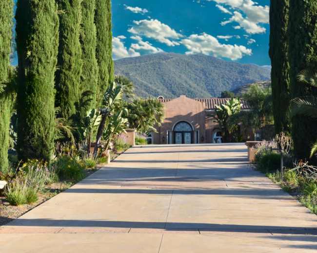 A long, paved driveway leads to a large, adobe-style house surrounded by tall cypress trees and lush vegetation, with mountains visible in the background under a partly cloudy sky.
