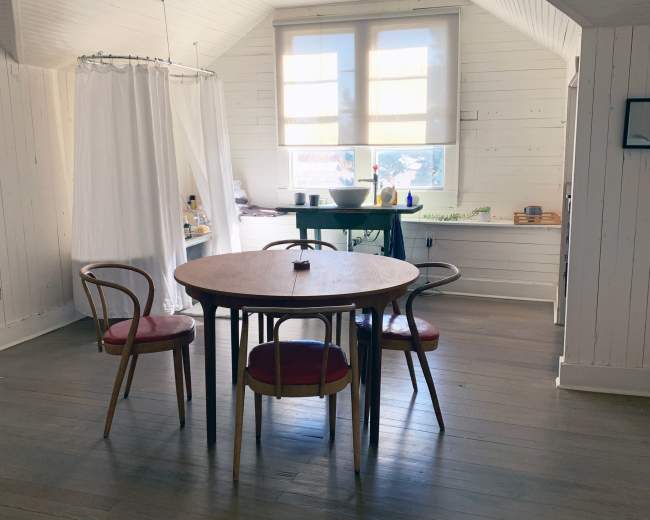 A wooden table with four chairs is situated in a room featuring white walls and large windows that allow natural light to enter.