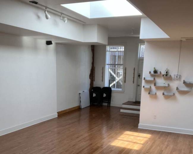 The image shows a spacious interior room with hardwood floors, a skylight, and minimal furnishings including two black chairs near the entrance.