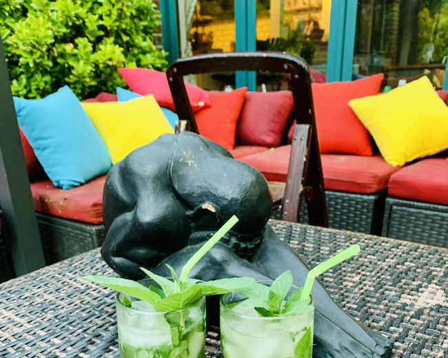 Two glasses of mojitos with mint leaves sit on a woven table beside a black sculpture of a reclining figure, with colorful cushions in the background.