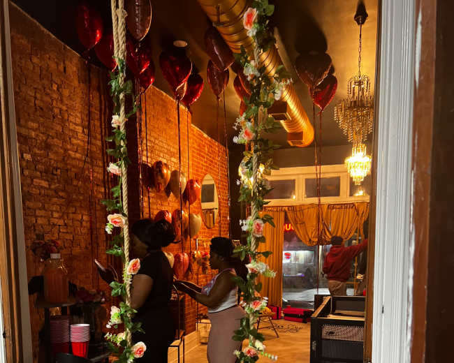 Two women stand near a swing adorned with flowers in a room featuring brick walls and decorative balloons.