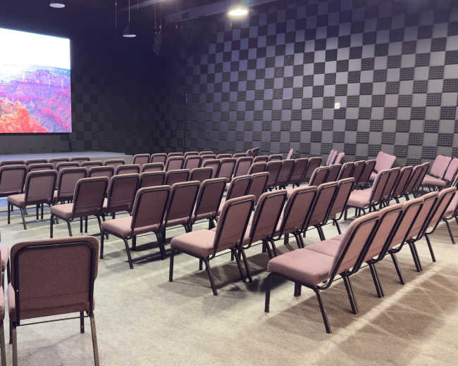 The image shows a conference room with rows of purple chairs facing a large projection screen and a patterned black wall.