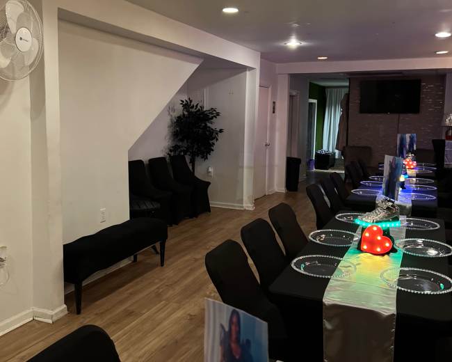 A long table set with elegant place settings occupies a well-lit dining area, flanked by black chairs and a decorative plant in the corner.