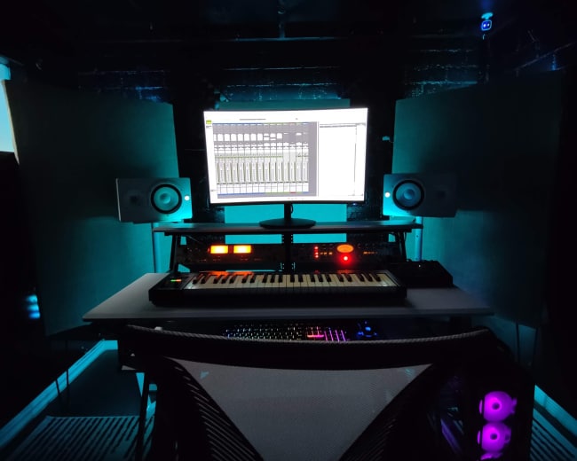 A music production studio features a computer monitor, a MIDI keyboard, and speaker monitors against a dark backdrop.