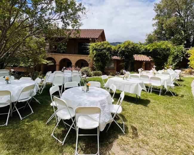 An outdoor event setup with round tables covered in white tablecloths arranged on a grassy area, surrounded by trees and a large building in the background.