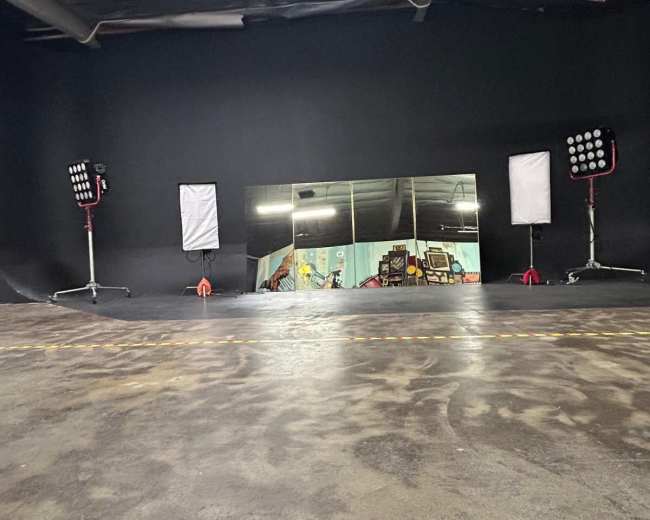 The image shows a dark studio space featuring several lights and a large mirror against a black wall.