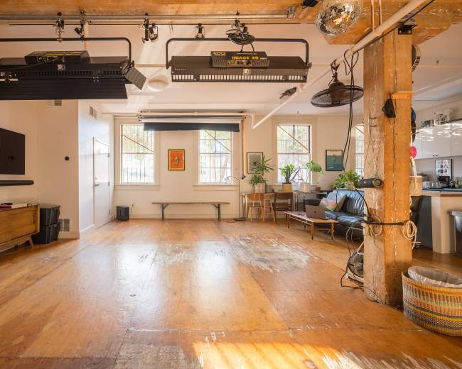An open room with wooden flooring, light fixtures mounted on the ceiling, potted plants, a black sofa, and a wooden bench along with a kitchen area visible in the background.