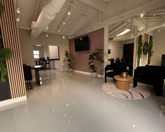 The image shows a modern, spacious office interior featuring a polished floor, seating areas, plants, and a wall-mounted television.