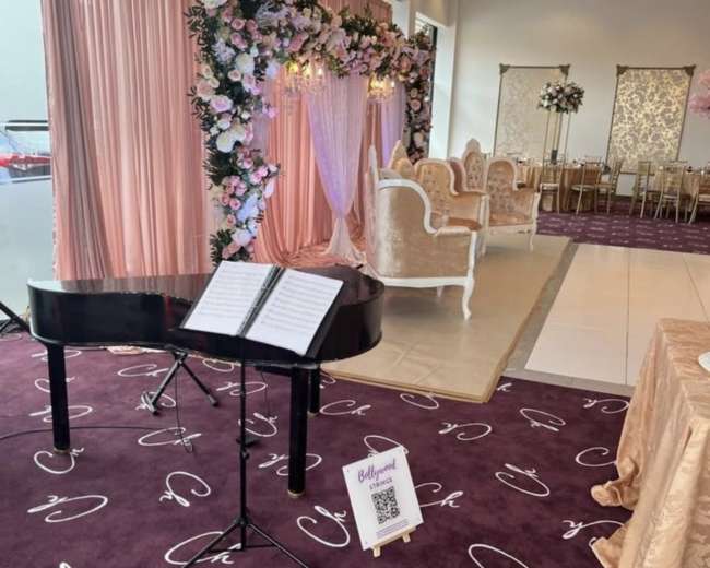 The image shows a decorated event space with a black piano and a backdrop of flowers, along with elegant seating arrangements in a light-colored setting.