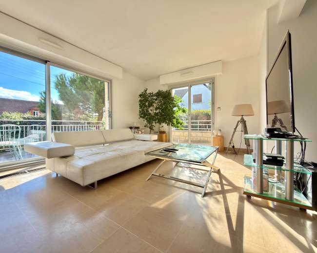 The image shows a bright living room with a white sectional sofa, a glass coffee table, and a television on a glass stand, alongside large windows that open to a balcony.