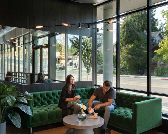 A man and woman sit on a green tufted sofa in a modern cafe with large windows, surrounded by plants and a round table.