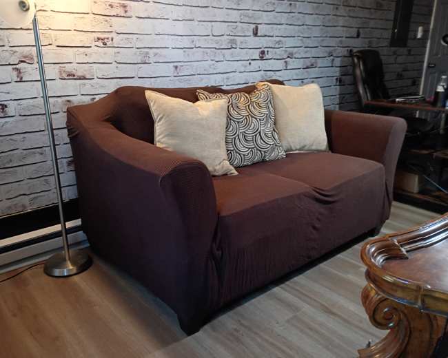 The image shows a brown sofa with two decorative pillows in front of a brick-patterned wall, alongside a standing lamp and a wooden table.