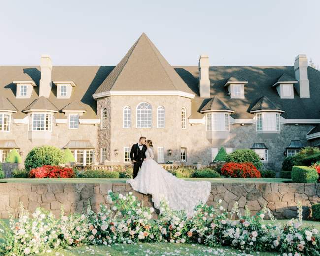 A couple in wedding attire stands together on a stone terrace in front of a large, intricately designed mansion surrounded by well-maintained flower beds.