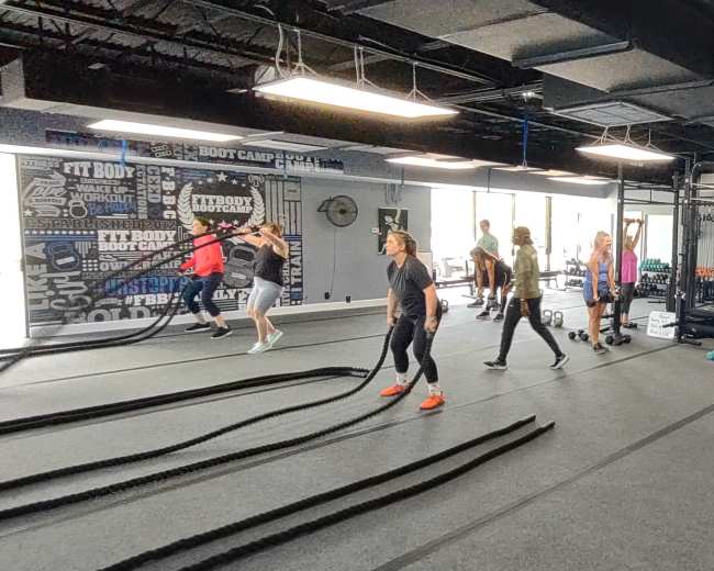 The image shows a fitness studio with several individuals engaged in various exercises, including using battle ropes and lifting weights.