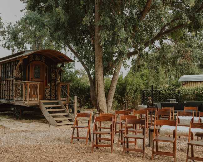 A charming wagon-style structure is positioned beside wooden chairs arranged for an outdoor gathering under large trees.