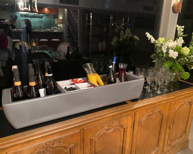 A modern bar setup features an ice-filled gray trough with various bottles of champagne, a pitcher, fresh berries, and an array of glasses, all displayed on an elegant wooden sideboard.