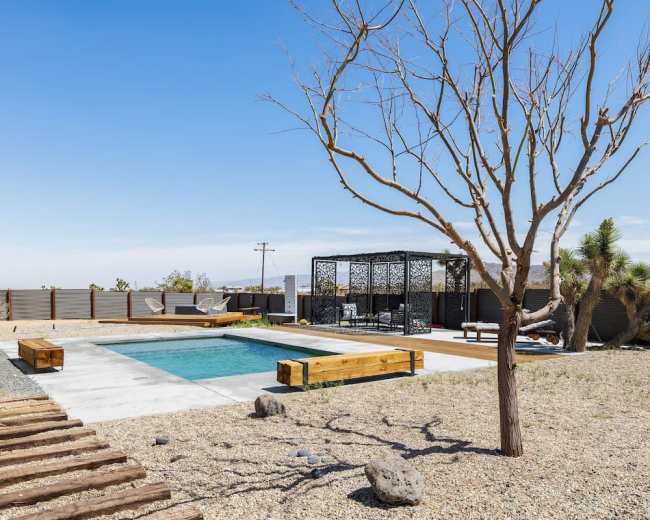 A desert landscape features a rectangular pool surrounded by gravel, wooden benches, and a shaded seating area under a gazebo.