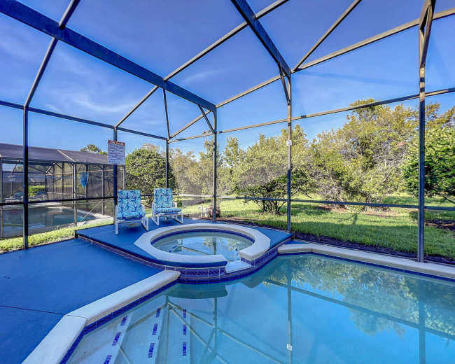 The image shows a screened-in pool area featuring a spa, two lounge chairs, and surrounding greenery.