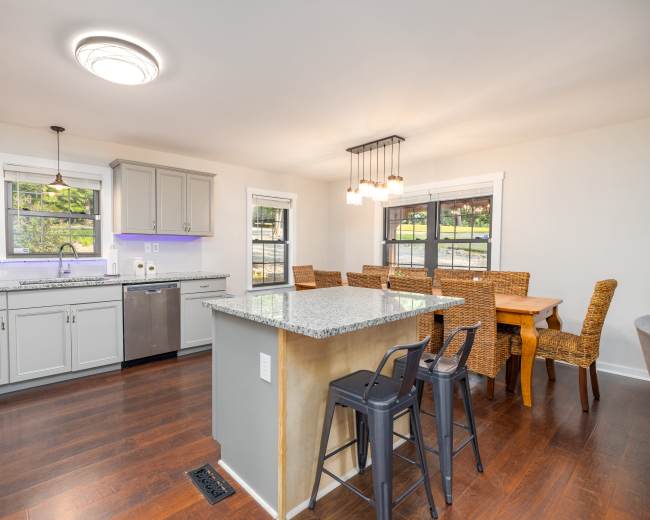 A modern kitchen features gray cabinets, a granite island, and a dining area with woven chairs and a wooden table.
