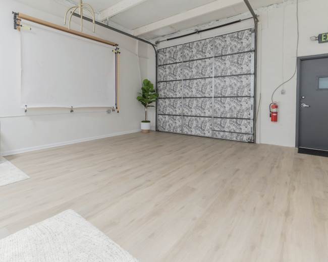 The image shows a spacious studio with light-colored wooden flooring, a white backdrop on a wall, a potted plant in the corner, and a large gray garage door.