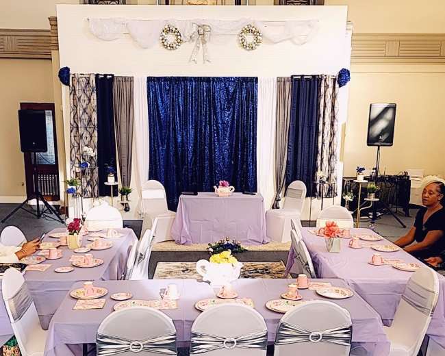 A banquet hall set up for an event, featuring tables with lavender tablecloths, decorative centerpieces, and a stage in the background with a blue curtain.