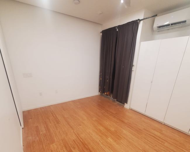 The image shows a sparsely furnished room with bamboo flooring, white walls, and a set of closed cabinets next to a window covered by dark curtains.
