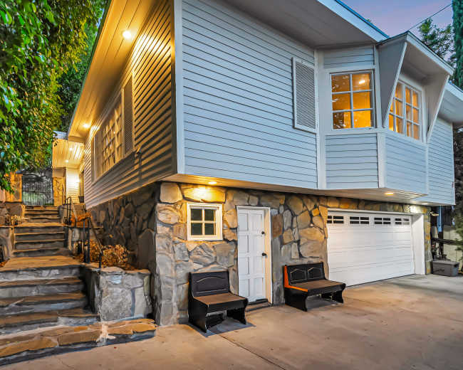 A modern house with a stone foundation has a set of stairs leading up to an entrance, and features a garage and two black benches in front.