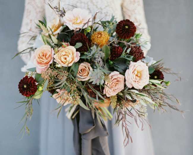 A person holding a bouquet filled with pink roses, brown dahlias, and greenery against a muted background.