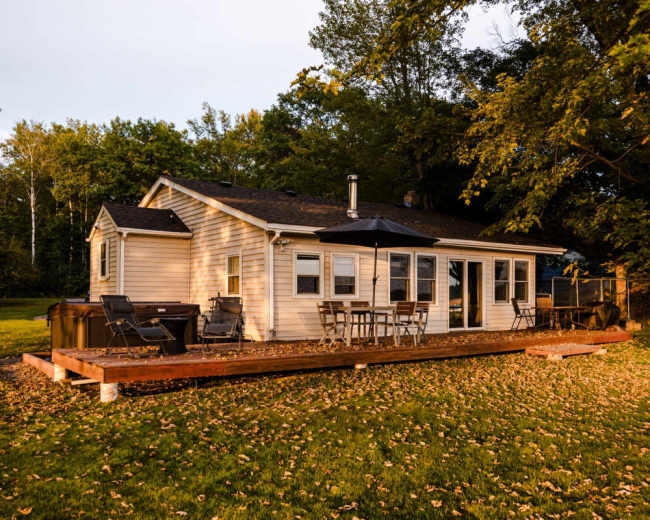 The image shows a single-story house with a wooden deck, surrounded by trees and fallen leaves.