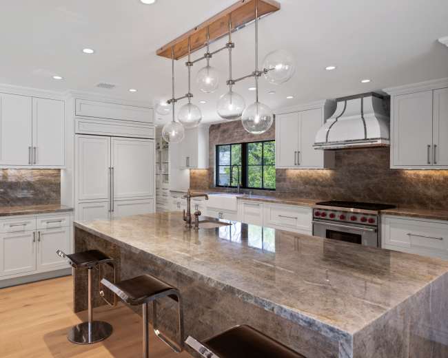 The image shows a modern kitchen with white cabinetry, a large gray marble island, and pendant lighting above.