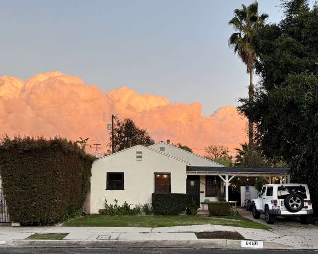 A single-story house with a white exterior, a front porch, and a Jeep parked on the driveway is situated under a colorful sunset sky.