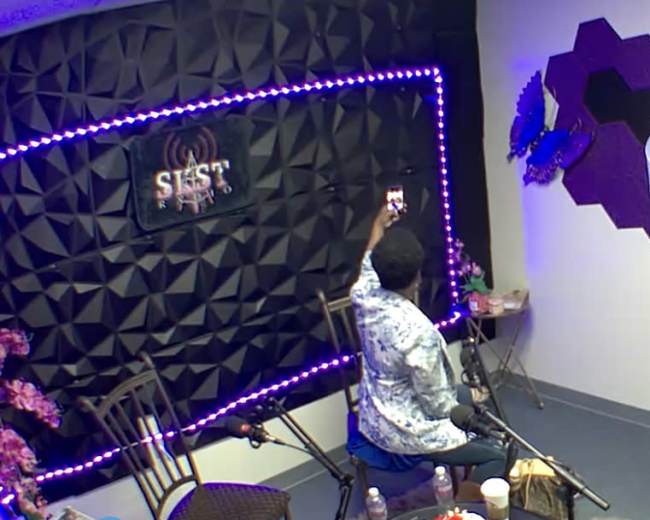 A person sits in a studio, holding a phone, with a backdrop of dim lighting and decorative elements on the wall.