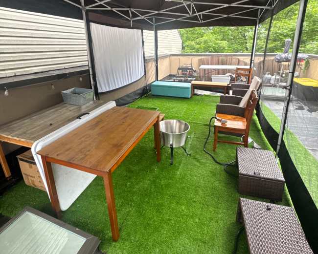 The image shows an outdoor tent setup with a green grass-like floor covering, several tables, chairs, and a small fire pit in a partially enclosed area.
