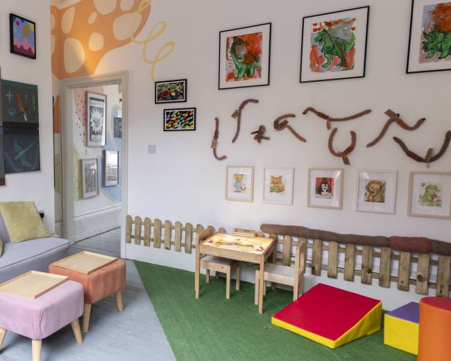 The room features a colorful play area with artwork on the walls, a small table surrounded by stools, and various seating arrangements.