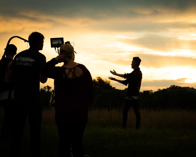 A group of people stands in a field as a person gestures towards the sunset in the background.