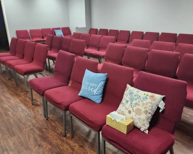 The image shows a room with rows of red chairs arranged for an event, featuring a decorative pillow and a small gift box on one of the chairs.
