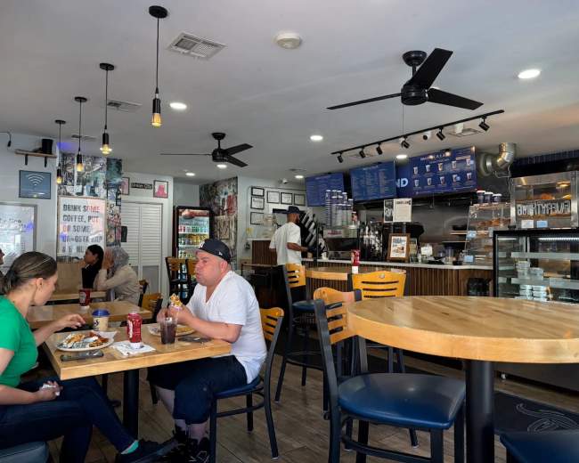 The image shows a casual café setting with patrons dining at tables while a barista prepares drinks at the counter.