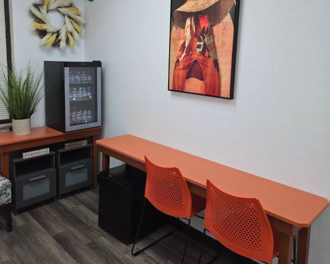 The image shows a small room with an orange table and chairs, a black mini fridge, and a framed artwork featuring a person in a hat on the wall.