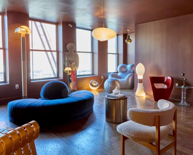 The room features a mix of modern furniture, including a curved blue sofa, red chairs, and unique lighting fixtures, all arranged around large windows.